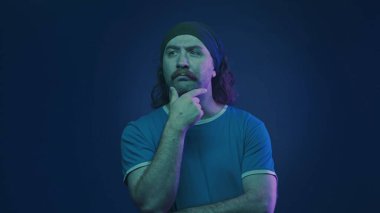 Stüdyoda saç bandı takan bıyıklı bir adamın portresi. Pembe ve yeşil neon ışıklı mavi arka planda. Bir adam bir şeyi dikkatlice düşünür, elini çenesine koyar.
