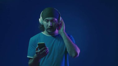 Beyaz kablosuz kulaklıklı bir adam, müzik de dahil olmak üzere akıllı telefonundaki bir çalma listesine göz atıyor. Stüdyoda, mavi arka planda, pembe ve yeşil neon ışıklı bir adam.