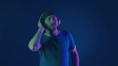 Beyaz kablosuz kulaklık takan bir adam müzik dinler ve dans eder. Stüdyoda, mavi arka planda, pembe ve yeşil neon ışıklı bir adam.