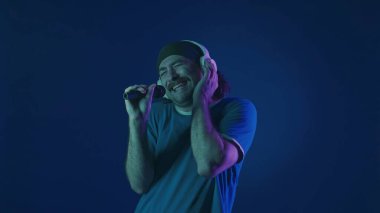 Beyaz kablosuz kulaklıklı bıyıklı bir adam dans ediyor ve mikrofona şarkı söylüyor. Stüdyoda mavi arka planda pembe ve yeşil neon ışıkları olan bir kadın var. Karaoke