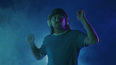 Kablosuz kulaklık takan bir adam müziğin tadını çıkarırken dans ediyor. Stüdyoda dans eden bir adam spot ışıkları altında, duman bulutlarında, pembe ve yeşil neon ışıklarda