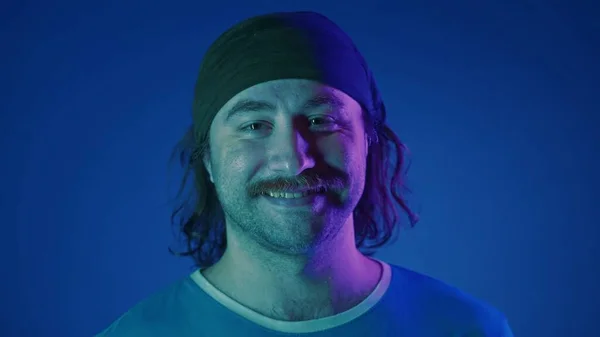 一个笑容可亲的长发男人的画像 头戴头巾 一个人在演播室里与蓝色背景的粉色和绿色霓虹灯相对照 — 图库照片