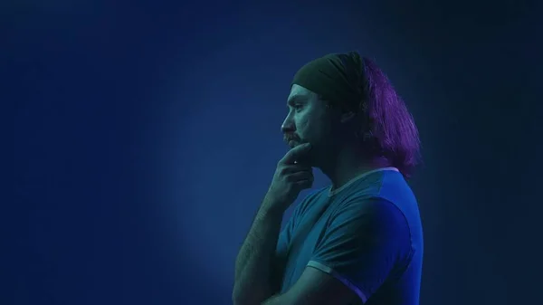 ピンクと緑のネオンライトの青い背景にスタジオでヘッドバンドを持つマッサージされた男性の側面のビュー 男は何かについて意図的に考えている 彼の顎に手を置く — ストック写真
