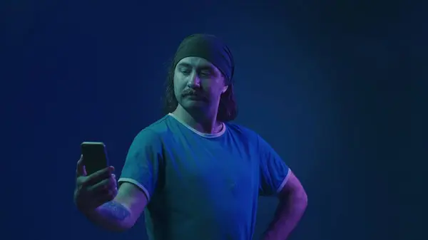 一个头带长发的大胡子男人手里拿着智能手机 摆出一副自私自利的架势 一个男人试图表现得有吸引力和性感 一个人在工作室里 蓝色背景 粉色和红色 — 图库照片