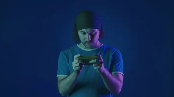 スマートフォンでオンラインゲームをプレイする緊張した男 ピンクと緑のネオンライトの青い背景にあるスタジオの男 — ストック写真