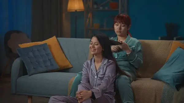 一张中等全景的照片拍摄到两个穿着睡衣坐在沙发上的年轻女性 她们一边谈论某件事 一边在做另一个人的头发 女孩之夜 过夜者 兄弟姐妹 — 图库照片