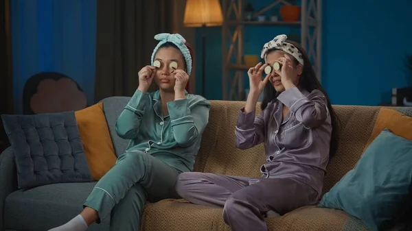 照片中 两个身穿睡衣的年轻女子坐在沙发上 眼前拿着黄瓜片 鬼鬼祟祟的 女孩之夜 过夜者 兄弟姐妹 — 图库照片