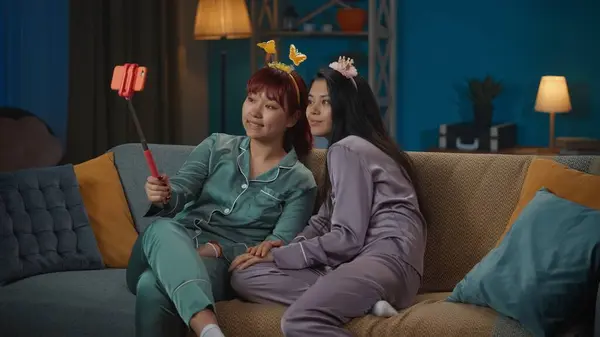 照片中 两个身穿睡衣的年轻女子坐在沙发上 用智能手机拍照 戴着可笑的头巾 女孩之夜 过夜者 兄弟姐妹 — 图库照片