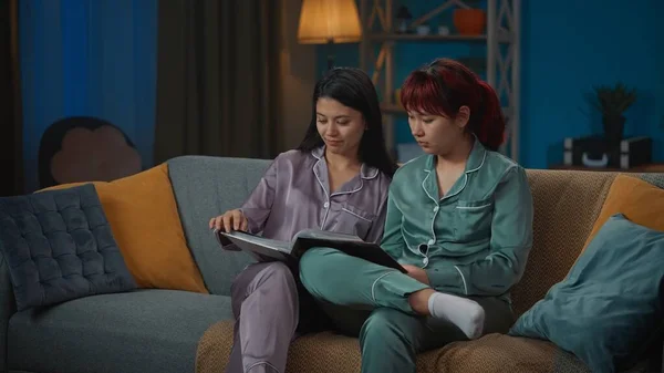 照片中 两个身穿睡衣的年轻女子坐在沙发上 翻阅着相册 回忆着她们的童年 聊着天 女孩之夜 过夜者 兄弟姐妹 — 图库照片