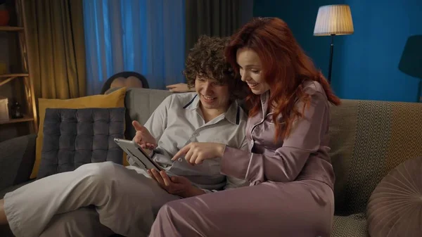 stock image Beauty and healthy relationships advertisement concept. Portrait of young couple spending time together. Man and woman in pajamas sitting on the sofa, holding tablet and watching something.