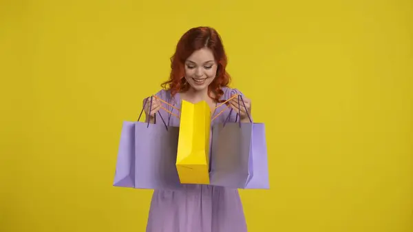 A redhaired woman with a satisfied expression looks into a shopping bag. Woman in the studio on a yellow background. Sale, Black Friday, advertising, discounts