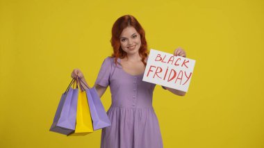 Elinde alışveriş poşetleri ve Kara Cuma yazan bir kadın var. Stüdyoda sarı arka planda kızıl saçlı çekici bir kadın var. Kara Cuma satış günü konsepti