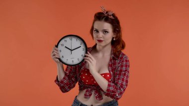 Çekimde kırmızı arka planda parlak renkli makyajlı genç kızıl saçlı bir kadın yer alıyor. Bir duvar saati tutuyor ve kameraya bakıyor. Bir beklentinin göstergesi. Orta boy.