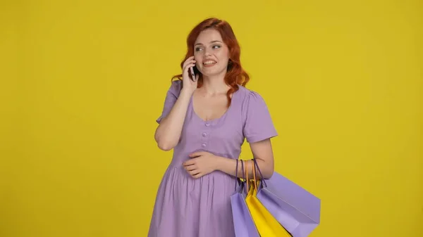 A woman with shopping bags speaks on a smartphone, shares her emotions from shopping. Redhaired woman in the studio on a yellow background. Sale, Black Friday, advertising, discounts
