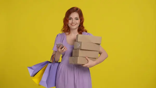 ショッピングバッグと紙箱を持つ若い女性は スマートフォンを使用しています 黄色の背景にあるスタジオの赤毛の女性 ブラックフライデー セール購入日のコンセプト — ストック写真