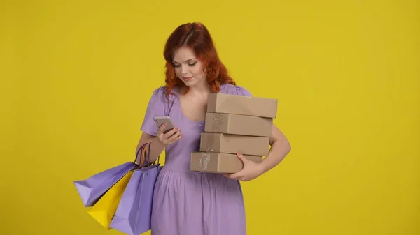 ショッピングバッグと紙箱を持つ若い女性は スマートフォンを使用しています 女性は有利なオファー プロモーションを通して見て オンライン注文を行います 黄色の背景にあるスタジオの赤毛の女性 — ストック写真