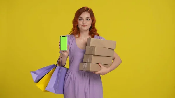 ショッピングバッグと紙箱を持つ若い女性は スマートフォンの緑色の画面を示しています 黄色の背景にあるスタジオの赤毛の女性 ブラックフライデー セールス コンセプト 広告エリア ワークスペース — ストック写真