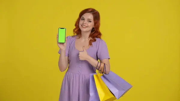 グリーンスクリーンでスマートフォンを保持しているショッピングバッグを持つ魅力的な女性と親指を表示します 黄色で孤立した若い女性 ブラックフライデー セールス コンセプト 広告エリア ワークスペース — ストック写真