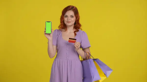 クレジットカードを持っているショッピングバッグを持つショッピングバッグとスマートフォンの緑色の画面を示すショップアホリック女性 黄色の背景にあるスタジオの女性 オンライン決済 広告エリア ワークスペース — ストック写真