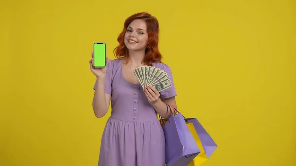 A woman with shopping bags holds a stack of dollar bills and shows a green screen of a smartphone. Redhaired shopaholic woman in studio on yellow background. Advertising area, workspace