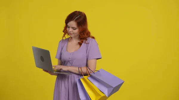 A woman with shopping bags is using a laptop, looking through great deals. Redhaired woman in a light sundress in the studio on a yellow background