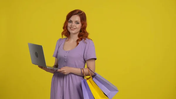 ショッピングバッグを持っている女性は 大きな取引を通して見て ラップトップを使用しています 黄色の背景にあるスタジオの明るい日没の赤毛の女性 — ストック写真