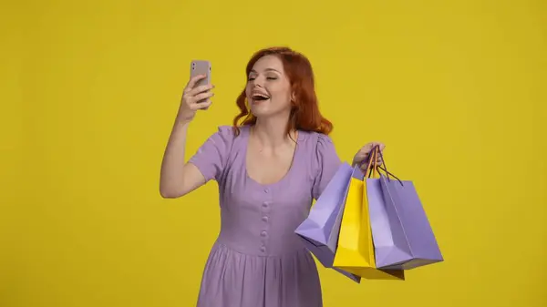 赤毛の女性はセルフィーを撮り スマートフォンを使ってビデオ通話をする 黄色の背景にスタジオでショッピングバッグをポーズする女性 — ストック写真