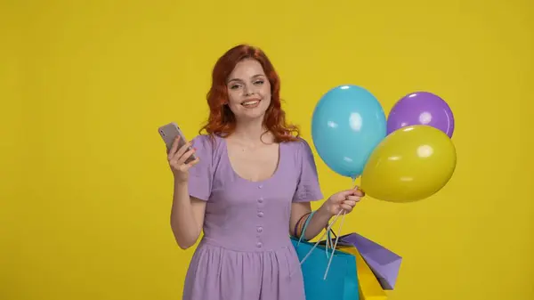黄色の背景にスタジオでショッピングバッグと風船を持つ女性 女性がスマートフォンを使用 — ストック写真