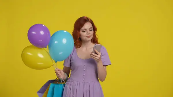 黄色の背景にスタジオでショッピングバッグと風船を持つ女性 女性がスマートフォンを使用 — ストック写真