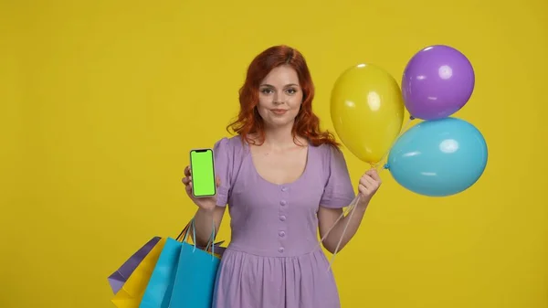 ショッピングバッグと風船を持つ若い女性は スマートフォンの緑色の画面を示しています 黄色の背景にあるスタジオの赤毛の女性 ブラックフライデー セールス コンセプト 広告エリア ワークスペース — ストック写真