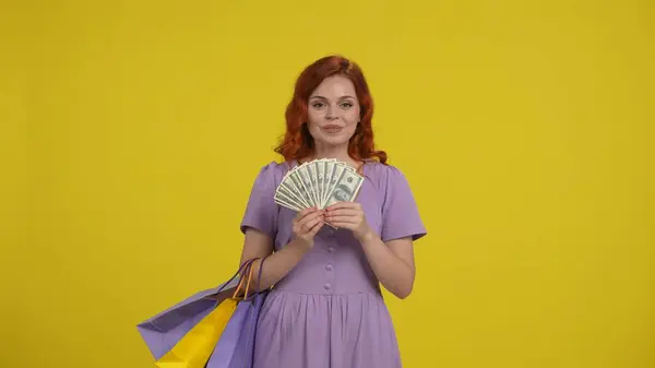 A woman with shopping bags holds a stack of dollar bills. Redhaired shopaholic woman in studio on yellow background. Advertising area, workspace