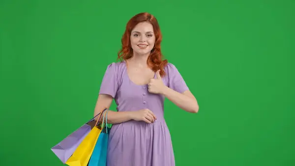 Redhaired woman with shopping bags shows a thumbs up. Woman in studio on green screen. Advertising area, workspace. Sale, Black Friday, discounts