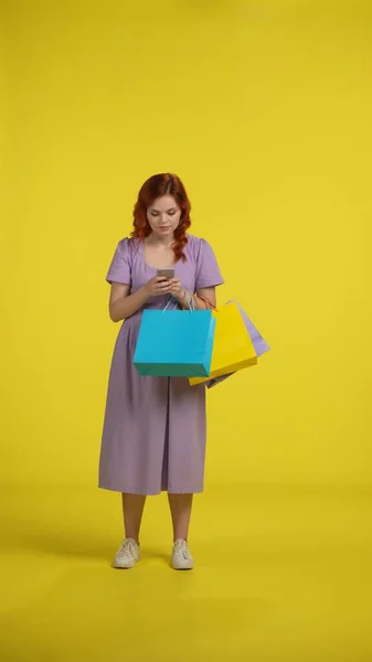 拿着购物袋的年轻女人用智能手机 一个女人浏览利润丰厚的工作机会 促销活动 并在网上下订单 全长红头发女人在工作室的黄色背景 垂直射击 — 图库照片