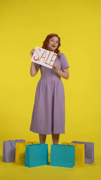 一位年轻貌美的女士站在购物袋周围 举着一面写着 的横幅 一个红头发的女人站在一间有黄色背景的演播室里 垂直射击 — 图库照片