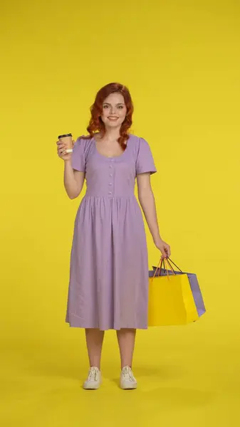 赤毛の女性は ショッピングバッグと紅茶またはコーヒーの紙コップを保持しています 黄色で隔離された全長日没の若い女性 垂直ショット 購入の割引を販売する概念 コピースペース — ストック写真