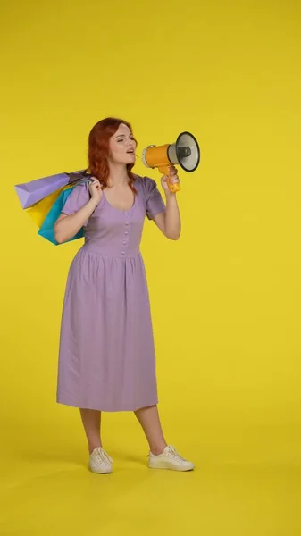 快乐的红头发女人 拿着购物袋对着扩音器说话 全长的女人在工作室的黄色背景 垂直射击 销售和折扣概念 — 图库照片
