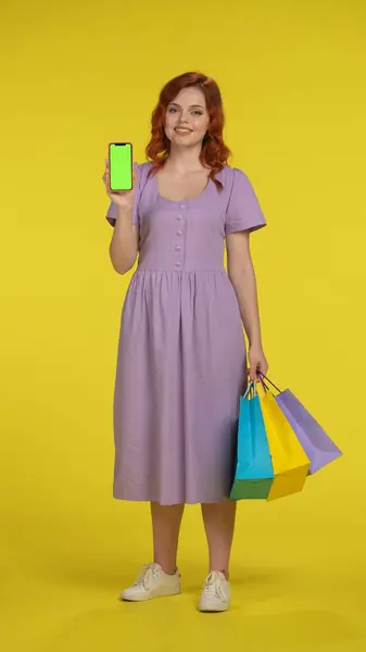 スマートフォンの緑色の画面を示すショッピングバッグを持つ若い女性 黄色い背景のスタジオで完全な長さのショッピングバッグを持つ赤毛の女性 垂直ショット ブラックフライデー セール購入日のコンセプト — ストック写真