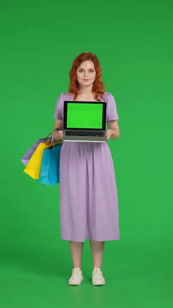 Redhaired woman holds a laptop with a green screen. Full length woman with shopping bags in studio on green screen. Vertical shot. Advertising area, workspace. Sale, Black Friday, discounts