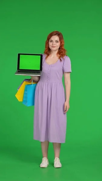 Redhaired woman holds a laptop with a green screen. Full length woman with shopping bags in studio on green screen. Vertical shot. Advertising area, workspace. Sale, Black Friday, discounts