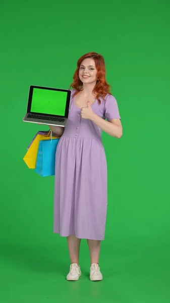 Redhaired woman holds a laptop with a green screen and showing thumbs up. Full length woman with shopping bags in studio on green screen. Vertical shot. Advertising area, workspace. Sale, Black Friday