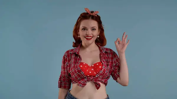 A young red-haired girl with bright makeup appears in the frame against a blue background. She is wearing retro clothes. She looks into the frame, smiles and shows a hand gesture Okay. Medium shot