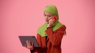 Orta ölçekli izole bir video, tesettür ve duvak takan çekici bir genç kadını çekiyor. Elinde bir tablet, elinde dizüstü bilgisayar, çalışıyor, telefonda konuşuyor. Kültürel çeşitlilik, iş ve eğitim.
