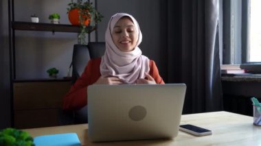 Orta ölçekli bir video, ofiste dizüstü bilgisayarın önünde oturan tesettürlü çekici genç bir kadının videosunu çekiyor ve heyecanlanıyor. Kültürel çeşitlilik, iş ve eğitim, reklam.
