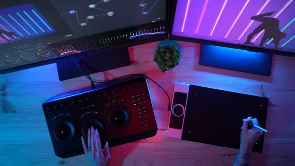 Video and photo editing advertisement concept. Freelancer working from home. Top view of woman hands working on pc in neon lights, editing photos and videos with graphic tablet and mixer board.