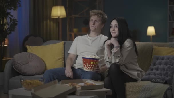 最初の愛と関係創造的なコンセプト 十代のカップルの肖像画 ソファーに座ってポップコーンを食べ 映画を見ている少年と少女 少年は恥ずかしがり屋の肩に手を置く 少女は抱擁を受け入れる — ストック動画