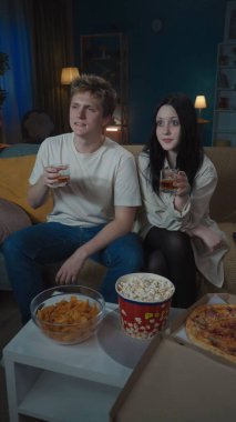 İlk ilişkiler yaratıcı konsept. Boş zamanlarını geçiren genç bir çiftin portresi. Kız ve erkek kanepede oturuyor, ellerinde viski bardaklarıyla film izliyorlar. Dikey fotoğraf.