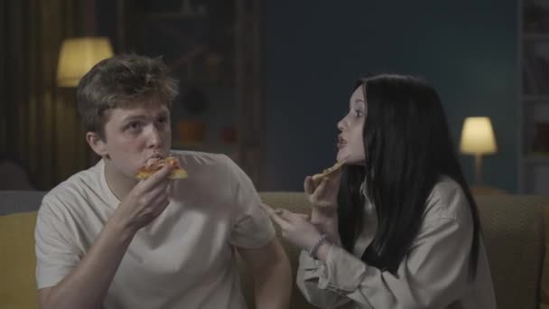 最初の愛と関係創造的なコンセプト 十代のカップルの肖像画は一緒に時間を過ごしています ソファーに座ってピザを食べたり 何かについて活発に話したり 笑顔で冗談を言ったりする少年と少女 — ストック動画