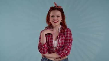 Çerçevede, mavi arka planda kızıl saçlı genç bir kadın var. O, kameraya bakıyor. O, eli ağzını kapatarak şakacı bir şekilde gülümser. Flörtleşmeyi gösteriyor, çok tatlı..