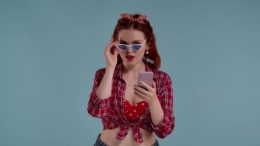 Mavi arkaplanda parlak makyajlı kızıl saçlı genç bir kadın var. Güneş gözlüğü takıyor, web sitelerine bakıyor ve telefonunda sosyal medya var. Gülümsüyor ve çok dikkatli.