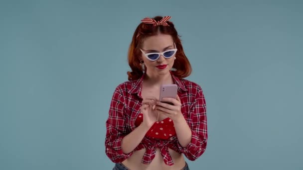 赤毛の若い女性 彼女は 彼女の携帯電話のウェブサイト ソーシャルメディアを通して見てフリップするサングラスを身に着けています その後 彼女は意図的に彼女のサングラスを上げてカメラに向かって見ます — ストック動画